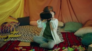Genç ve sevimli bir çocuk kapalı, rahat ve modern görünümlü bir çadırda siyah VR kulaklığıyla oynuyor ve havaya bir şeyler doğrultuyor. 4k