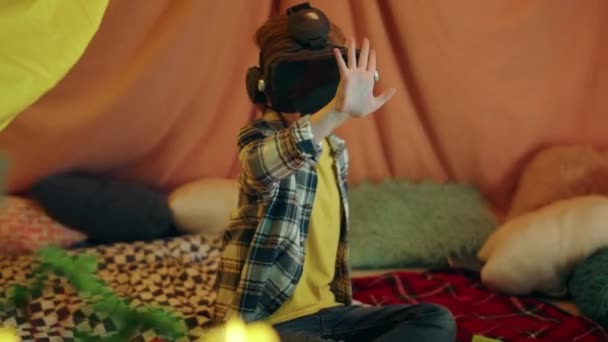 幼い少年は 自宅で作られた屋内テントの中で大量の枕や毛布に囲まれている間 顔に仮想現実の頭をセットして好奇心と愛らしい遊びをしています — ストック動画