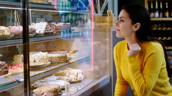 Taking details in the bakery cafe of a beautiful woman customer looking at the showcase fridge full of fresh deserts to choose the favourite one.