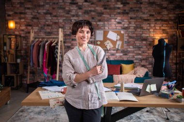 Modern terzisindeki karizmatik ve çekici terzi kadının önünde poz vererek terzi makasını tutuyor ve tatlı tatlı gülümsüyor..