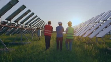 Fotovoltaik güneş panellerinin ortasında gün batımında yürüyen ekoloji mühendisleri grubunda güneş panellerinin temizliği ve güneş sisteminin işleyişi hakkında tartışıyorlar..
