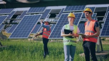 Güneş enerjisi çiftliğinde güvenlik üniforması içinde duran ve güneş ve panellerin temizliğini inceleyen bir grup teknisyen. 4k