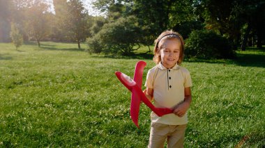 Parkta, güneşli bir günde kameranın önünde kırmızı bir uçağı tutarken kameraya doğru gülümseyen sevimli küçük bir çocuk. Portre