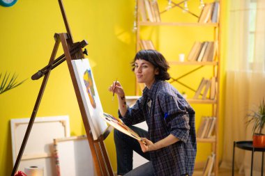 Bir kadın ressam, tuvale modern sanatsal ve soyut bir tablo çiziyor ve bunu yaparken çok neşeli görünüyor..