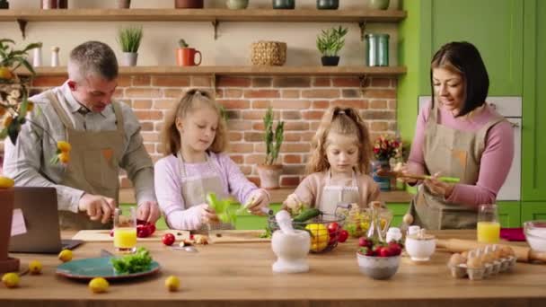 家庭のモダンなキッチンで白人の両親と彼らの2人の子供たちは一緒に夕食の準備を喜んで健康的なサラダのためにいくつかの野菜を切りました — ストック動画