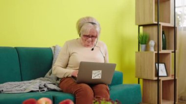 Oturma odasındaki kanepede dizüstü bilgisayarını internet kursları için kullanan yaşlı kadın kanepede oturuyor ve dizüstü bilgisayarına bakıyor ve kulaklığı takıyor. ARRI Alexa Mini 'de çekilen