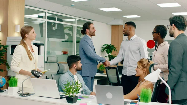 A boss woman in a white suit is talking to everyone in the meeting and smiling in a large modern office.