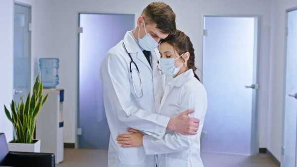 在明亮的医院走廊的M处 一对戴着防护面具的医生在摄像机前互相拥抱着 他们的脸很忧郁 肖像画 — 图库照片