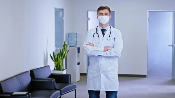 在一个现代化的医院走廊里 站在摄像机前的是一位严肃的医生 他双手交叉 直勾勾地看着摄像机 戴着防护面具 肖像画 — 图库照片