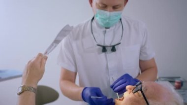 Kameraya yakın, beyaz görünümlü, sağlık kontrolü yapan doktor dişçi sandalyesinde oturan ve çürüklere bakmak için ayna tutan yaşlı bir adamın dişlerini inceliyor. 4k