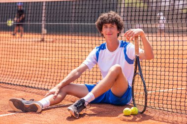 Tenis takımının yanında profesyonel tenisçi, dışarıda tenis kortunun zemininde dinlenirken kameraya bakıyor..