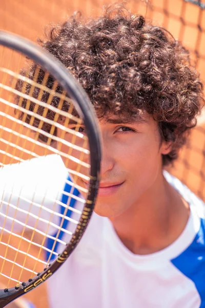 Closeup capturing video of a charismatic young tennis player he sitting down on the clay court beside the tennis net and playing with the racket looking to the camera. Portrait