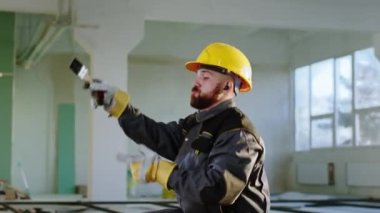 Mutlu ve karizmatik inşaat işçisi şantiyede güvenlik kaskı ve özel üniforma giyerek ve kablosuz kulaklıklardan müzik dinleyerek eğleniyor. 4k
