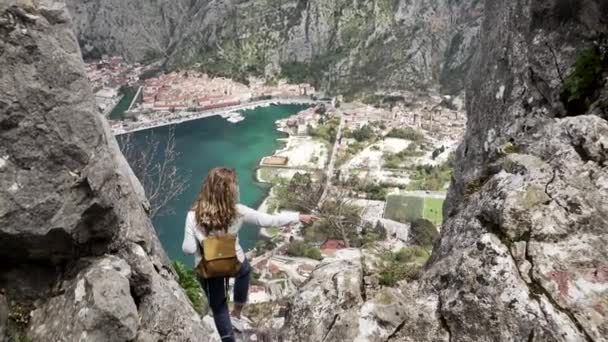 Uma menina turística com uma mochila fica no topo de uma montanha e olha para a cidade — Vídeo de Stock