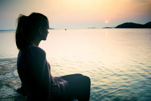 Mujer joven sentada en la playa al atardecer, símbolo de sueños y anhelo —  Fotos de Stock