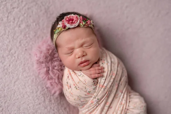 Little newborn Ukrainian girl sleeps peacefully on a pink background. On his head a bandage of flowers.