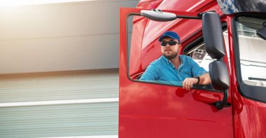 Beyaz tenli yarı kamyon şoförü. Araç kabininde güneş gözlüğü takıyor. Uzun bir yolculuğa hazırlanıyorum. Ağır Ulaşım Endüstrisi TemasıName.