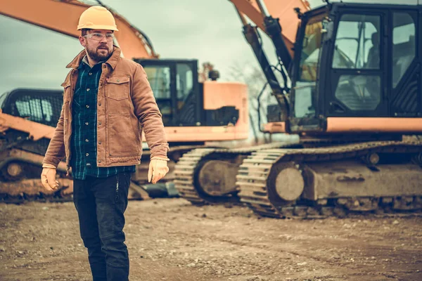 黄色のハード帽子をかぶって掘削機のような重機の間に滞在する30代の地上作業監督者 — ストック写真
