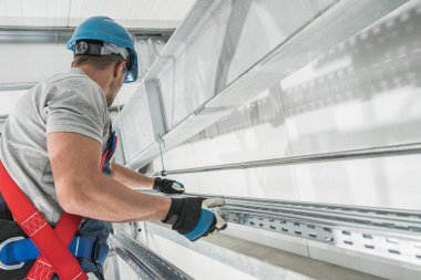 Güvenlik kemeri takan inşaat işçisi ve yeni geliştirilmiş bir ticari depo binasında çalışan sert şapkalı alüminyum elementleri montaj için hazırlıyor..