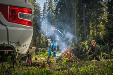 Caucasian Friends in Their 40s Hanging Out Next to Campfire and RV Camper Van in Scenic Woodland Wilderness. Recreational Vehicle Dry Campsite. clipart