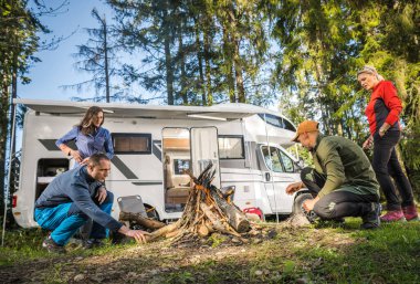 RV Road Trip Vacation with Friends. Two Caucasian Couples Hanging Around Campfire Next to Their Motorhome Camper Van. Dry Camping in a Forest.