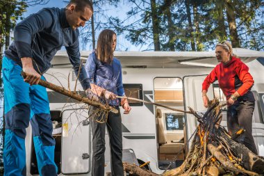 Three Caucasian Friends Hanging Out Around Campfire Next to Their Motorhome Recreational Vehicle. Summer Weekend Getaway Theme.