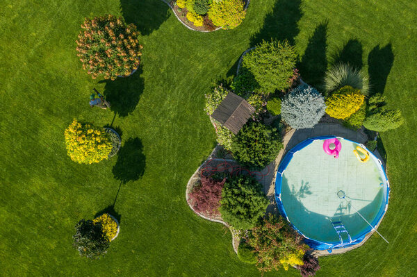 Beautifully Landscaped Garden at the Backyard with Swimming Pool and Perfectly Maintained Lawn. Professional Gardener at Work Taking Care of Plants. Hot Summer Day with Trees Casting Shadows on the Grass. Aerial View.