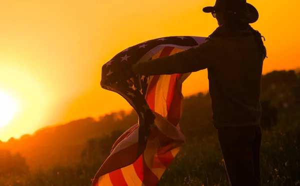 欧米風の男性は手にアメリカ風の旗を持っている 美しい夕日を背景にシルエット 愛国的テーマ — ストック写真