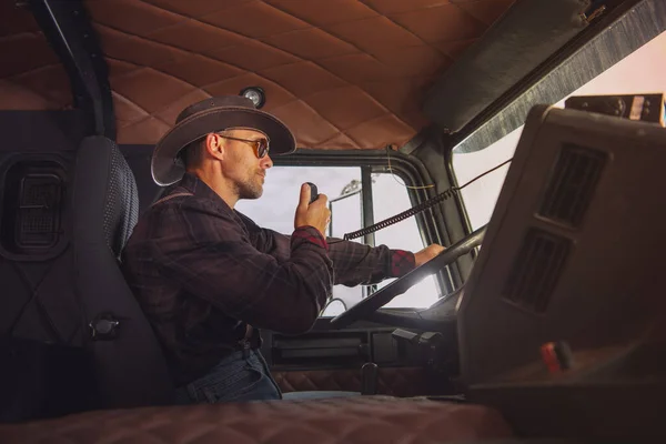 Caucasian Trucker Wearing Western Style Outfit Driving His Semi Truck and Talking Through CB Radio. Side View.