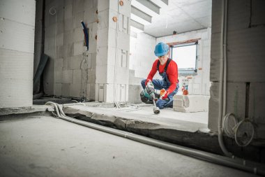 Güvenlik Miğferli Beyaz İnşaat İşçisi ve Gözlüklü Delik Testere Bıçak Eklentisini Güç Matkabıyla Kullanarak İşini Yapıyor. İnşaat alanı arka planı. Gayrimenkul Geliştirme Teması.