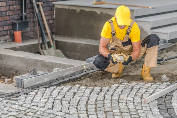 Caucasian Contractor in His Work Wear Carefully Laying Granite Setts in His Client\'s Front Yard. Landscaping of the Area Around a Newly Constructed Residential Building. Construction Theme.