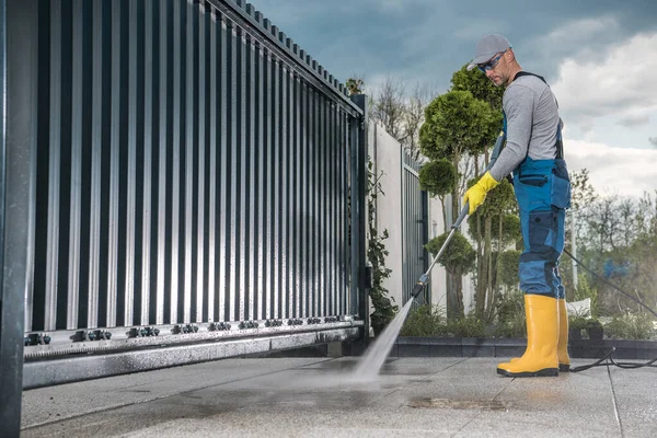 仕事中の白人労働者職業用圧力洗浄機を使用して入口ゲートの前でコンクリートドライブウェイを掃除する黄色のラバーブーツ プロパティメンテナンスのテーマ — ストック写真
