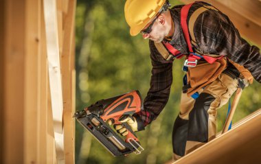Caucasian Construction Worker with a Nail Gun Assembling Wooden Skeleton Frame Beams in a Newly Developed Building. Construction Industry Theme.