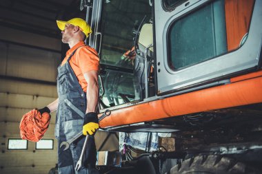 Heavy Duty Industrial Equipment Machinery Maintenance and Repair Service. Caucasian Worker with Wrenches in His Hand Proudly Standing Next to Freshly Fixed Digger Machine in His Workshop.