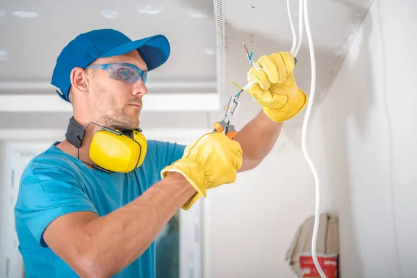 Closeup of Caucasian Electrician in Protective Gloves and Glasses Doing Illumination Installation Work Stripping Cables with Professional Wire Stripper. Residential Building Renovation Work Theme.