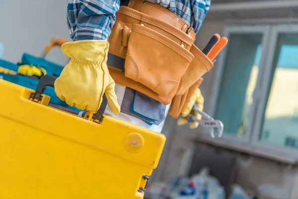 Closeup of Yellow Toolbox Carried by Construction Worker Wearing Leather Tool Belt and Protective Gloves. Building Site in the Background.