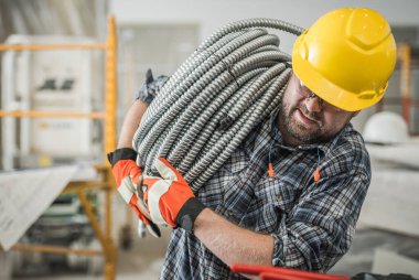 Closeup of Caucasian Contractor in Yellow Safety Helmet Putting Efforts to Move Heavy Metal Electric Cables Conduit Pipe Carrying Them on His Shoulder. Construction Work Theme.