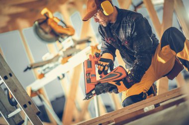 Caucasian Construction Worker in Safety Headphones Building a Wooden Frame of Canadian Style Residential House Using Nail Gun Tool. Industrial Theme.