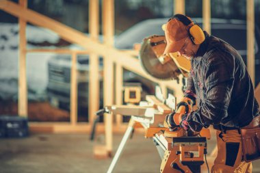 Caucasian Contractor Working with Wood While Constructing the Wooden Frame of Canadian Style Residential Building. Construction site in the Background.