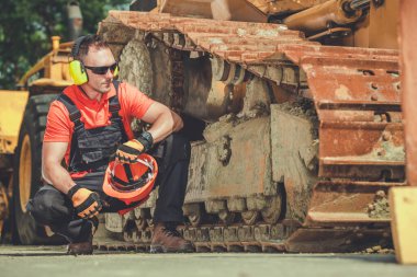 Caucasian Construction Worker Wearing Noise Reduction Headphones and Holding Red Hard Hat in His Hand Resting in Front of His Heavy Industrial Equipment Machine.
