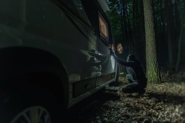 Man Fully Dressed in Black With Covered Face Trying to Break Into a Camper Van Parked in the Woods at Night. Robbery During Traveling. Criminal Theme.