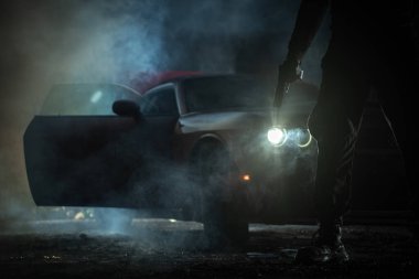 Silhouette of the Gangster with a Gun in His Hand Standing in Front of His Car in the Darkness. Criminal World at Night.