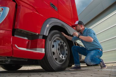 Caucasian Trucker Closely Checking the Condition of the Tires in His Large Heavy Duty Red Semi Truck Before Setting Off to Deliver a Cargo. Transportation and Logistics Workers Theme.