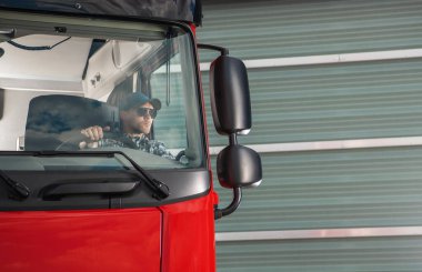 Caucasian Truck Driver in Sunglasses and Baseball Cap Driving His Red Semi Truck Out of the Logistics Center Car Park Fully Loaded and Ready to Start Delivering His Cargo.