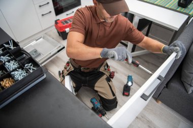 Caucasian Male Worker Installing the Handle to the Kitchen Cabinet Door Using Screwdriver. Modern New Kitchen and Other Professional Tools for Installing and Repairing Furniture in the Background.