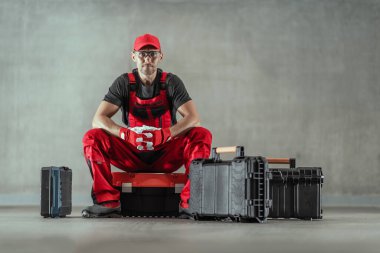 Caucasian Handyman in Red Work Uniform Sitting on the Toolbox Surrounded by More Boxes of His Professional Repair Tools. Solid Grey Background. Horizontal Photo.