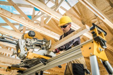 Construction Worker in Yellow Hard Hat Cutting a Wooden Plank with Circular Saw Power Tool. Wooden House Building Site in the Background. Industrial Theme.