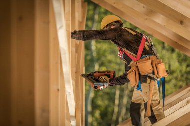 Caucasian Male Worker Wearing Safety Harness and Hard Hat on the Construction Site of Wooden Residential Building. Working in the Roof Section, Driving Nails Into Wood Planks Using Nail Gun.