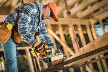 Caucasian Middle Aged Builder Working with Electric Wood Planer Power Tool at the Construction Site of Wooden Skeleton of Canadian Style Residential Building. Industrial Theme.