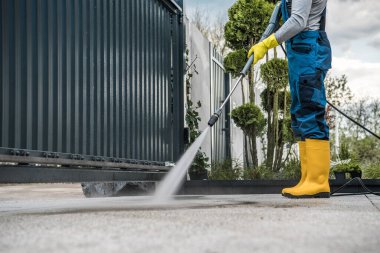 Worker Wearing Protective Uniform and Yellow Rubber Boots Cleaning the Driveway Behind the Entrance Gate Using Pressure Washer Machine. Yard Maintenance Theme.
